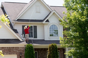 man cleaning the upper floor exterior of his home standing on the lower roof using a pressure sprayer | removing rush from vinyl siding
